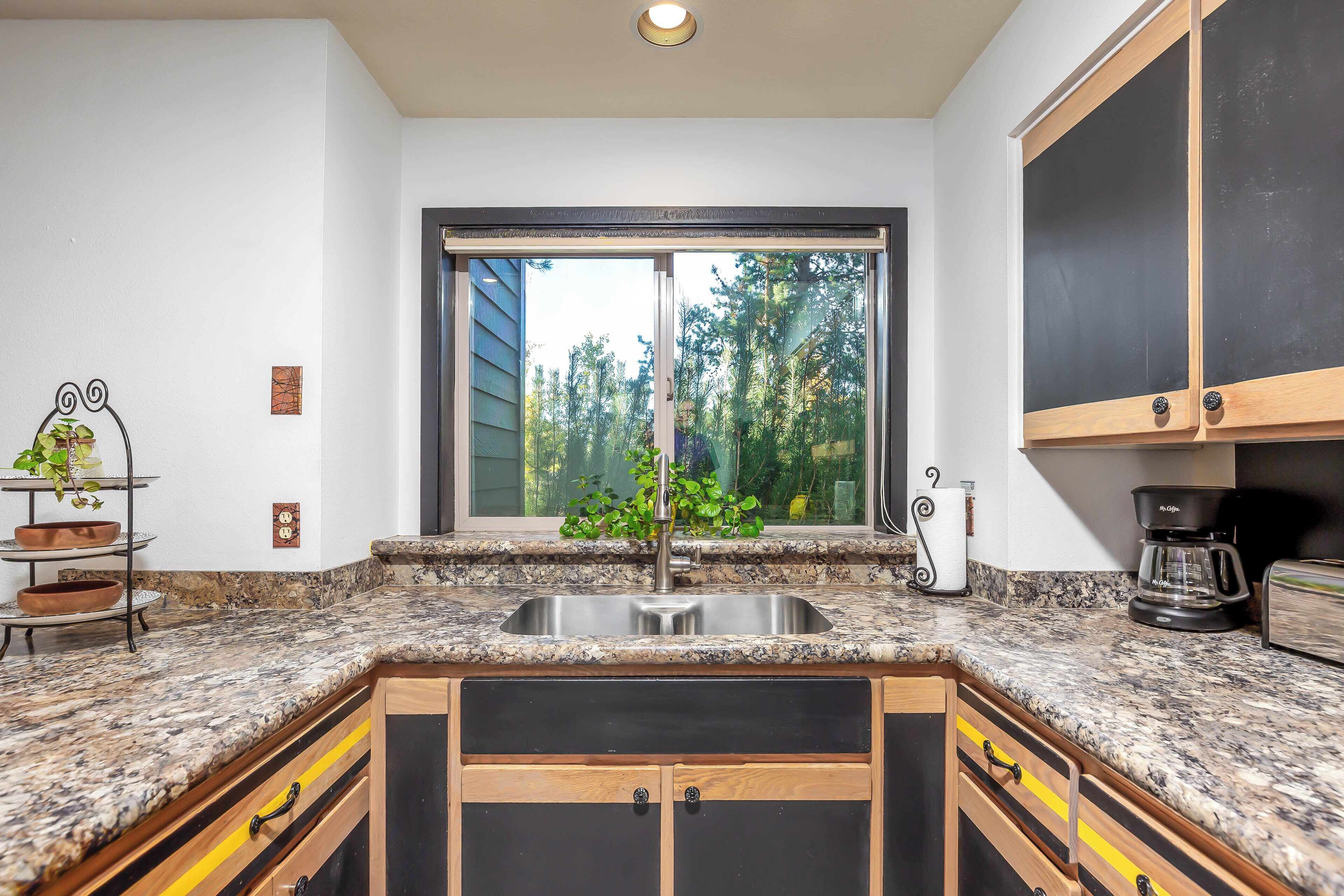 A kitchen with a granite countertop, a double sink, wooden cabinets, and a window with a view of trees.