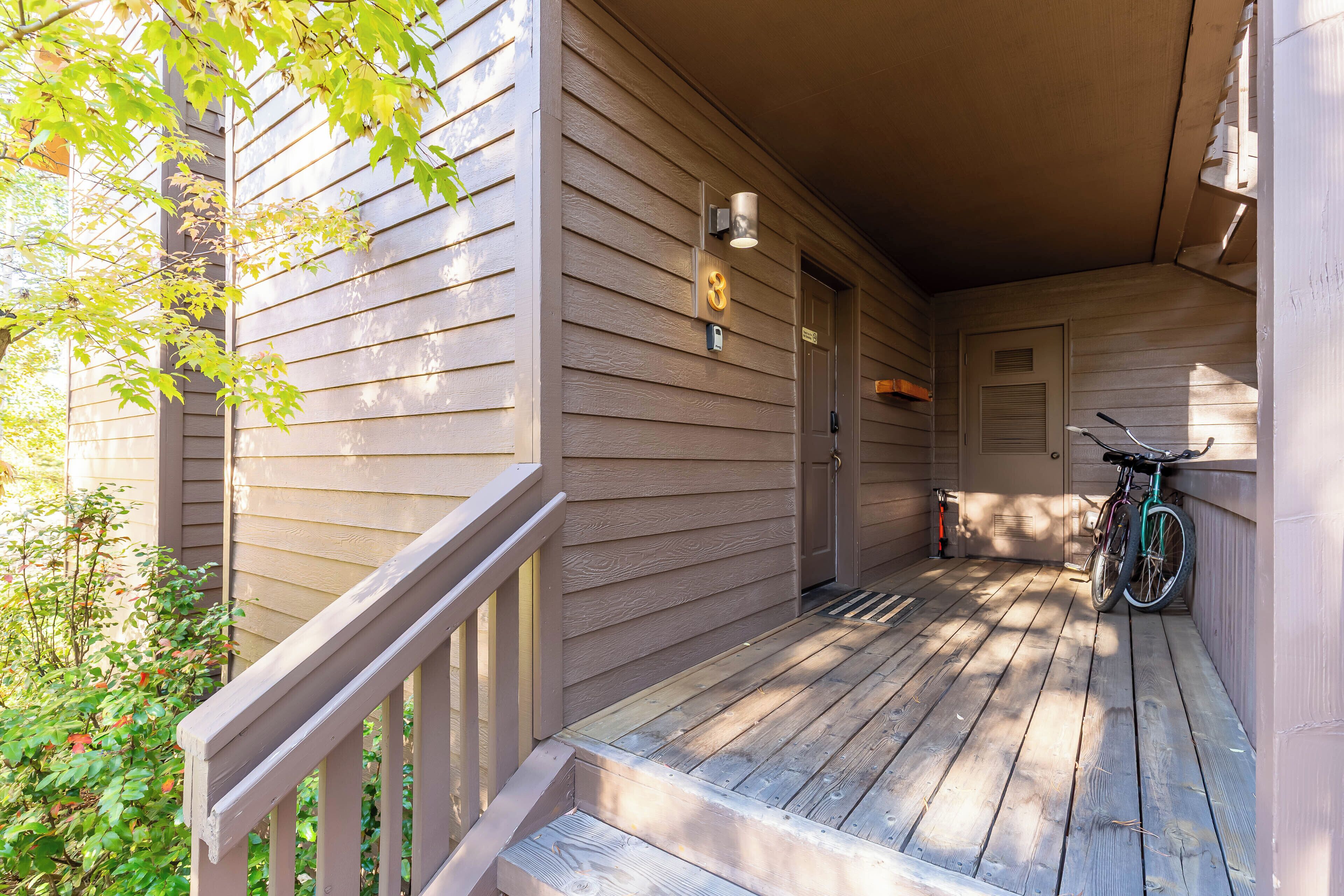 A wooden porch with a bicycle leaning against the wall, a door, and a small outdoor area with a bench.