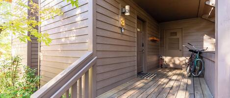 A wooden porch with a bicycle leaning against the wall, a door, and a small outdoor area with a bench.