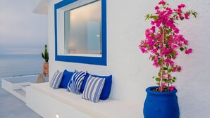 Pared exterior blanca con un ventanal de marco azul, una jardinera azul con flores rosadas y un banco blanco con cojines a rayas azules y blancas.