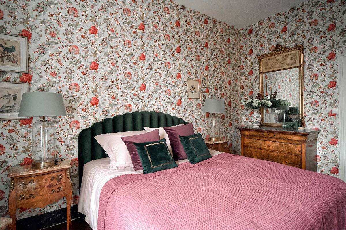 A bedroom with floral wallpaper, a bed with a green upholstered headboard, two bedside tables with lamps, and a dresser with a mirror.