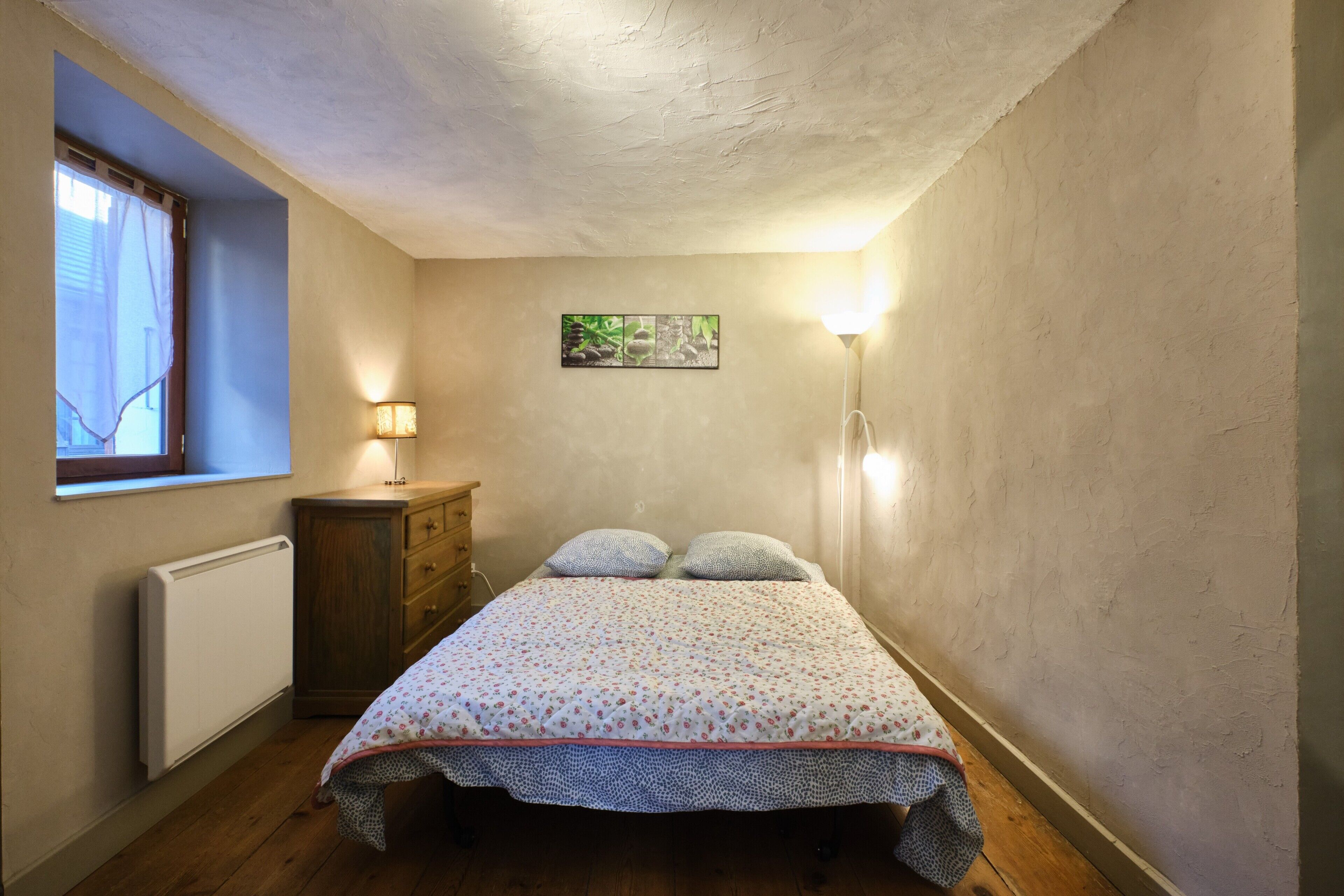 A bedroom with a bed, a dresser, a radiator, a window with curtains, and a wall-mounted picture.