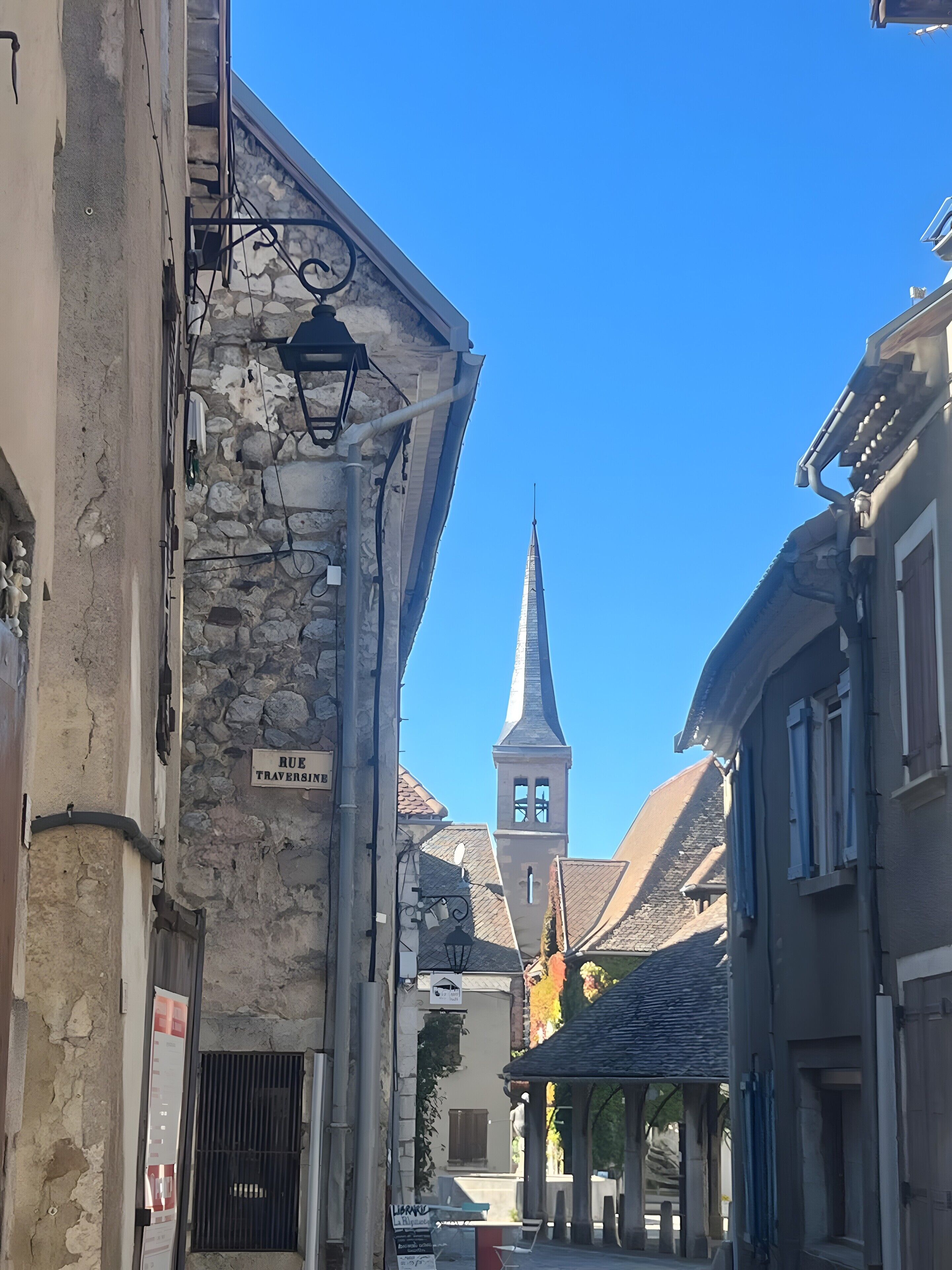 Une rue étroite bordée d’immeubles en pierre, avec un clocher d’église et une enseigne indiquant « RUE TRAVERSIN ».