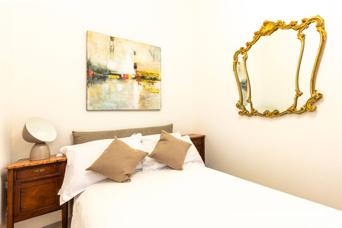 A neatly made bed with two pillows, a bedside lamp, a wooden nightstand, and a framed abstract painting on the wall.