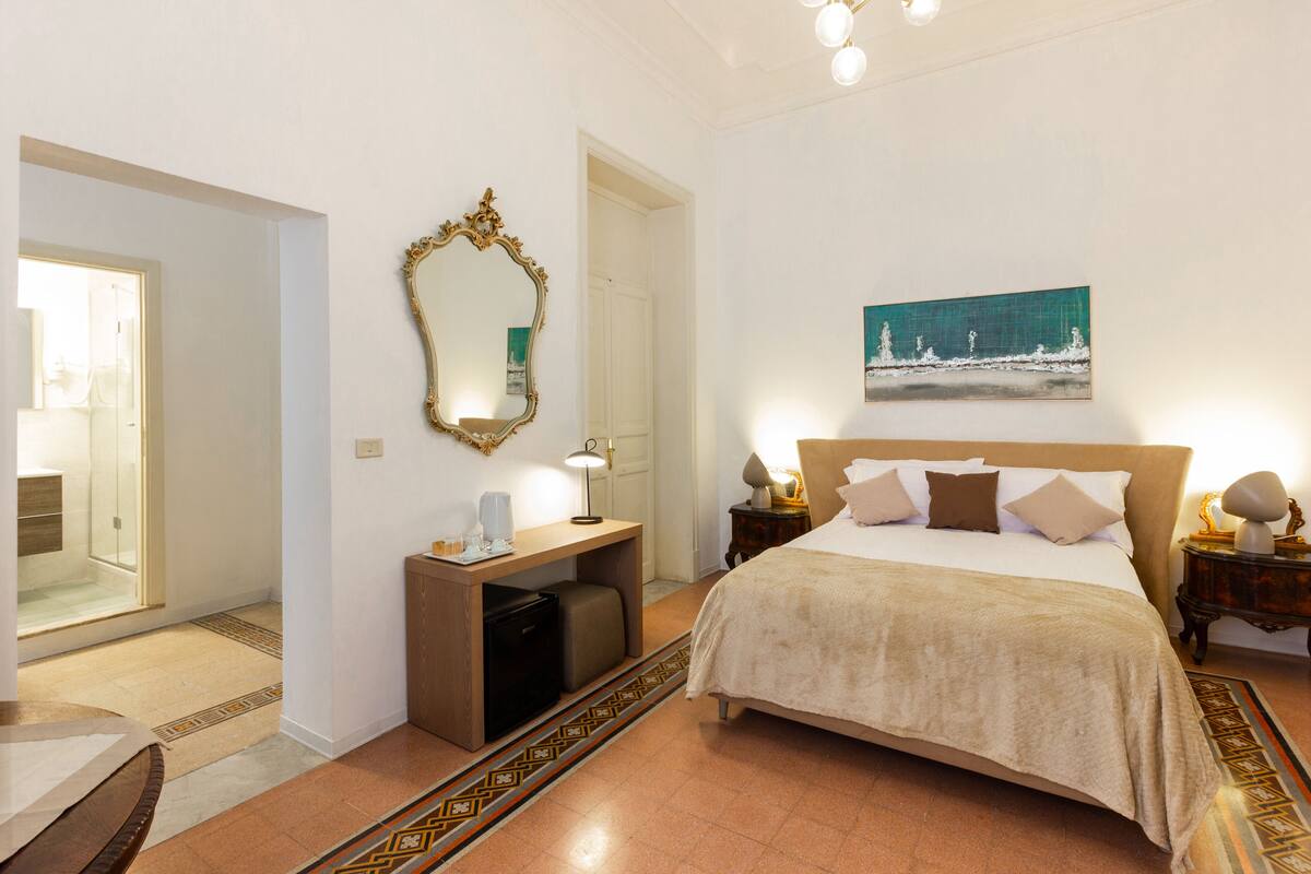 A hotel room with a bed, bedside tables, a desk, a mirror, and a painting on the wall.
