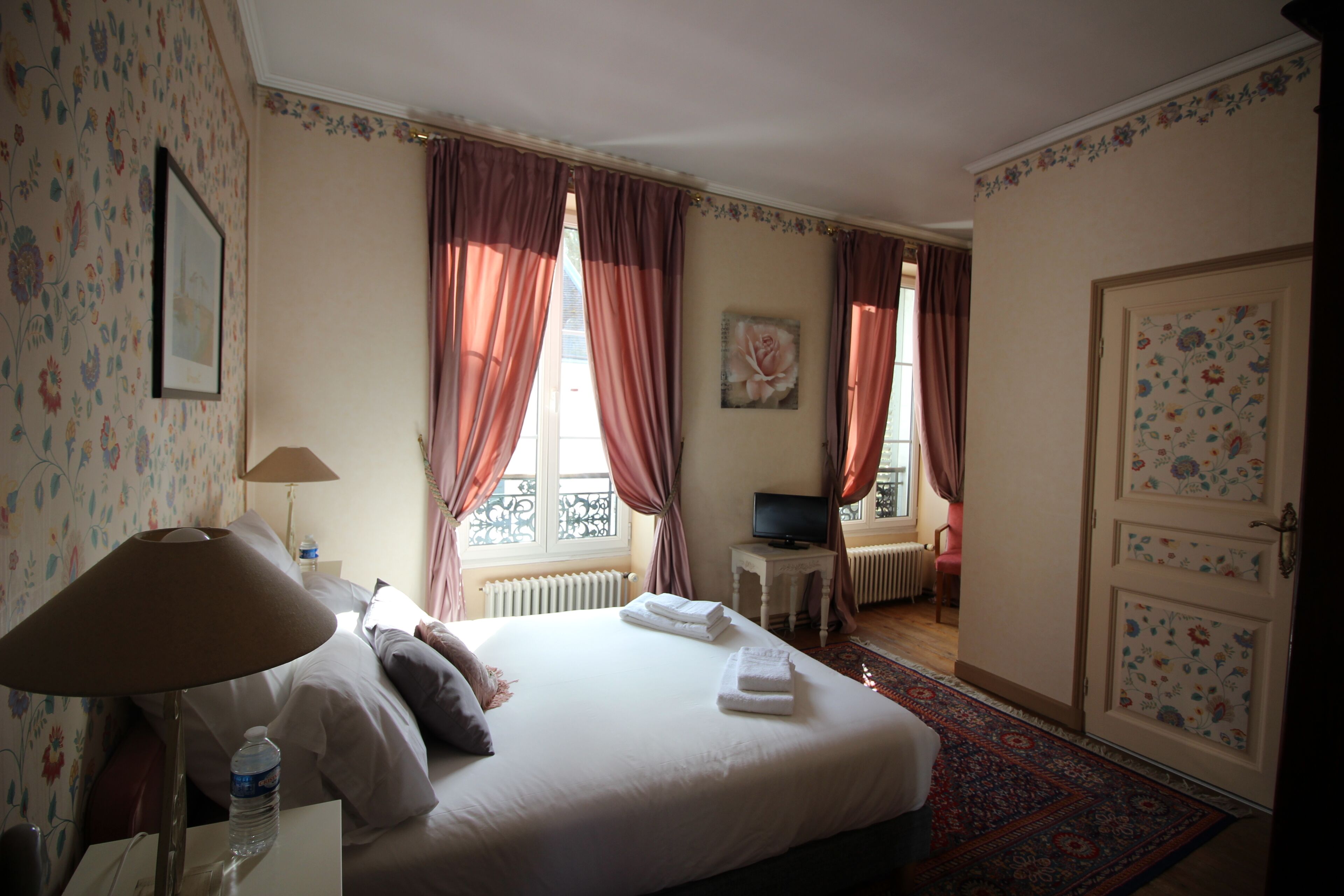 Ein Hotelzimmer mit einem Bett, einem Fernseher, einem Heizkörper, einem Fenster mit Vorhängen und einer Tür mit Blumenpaneelen.