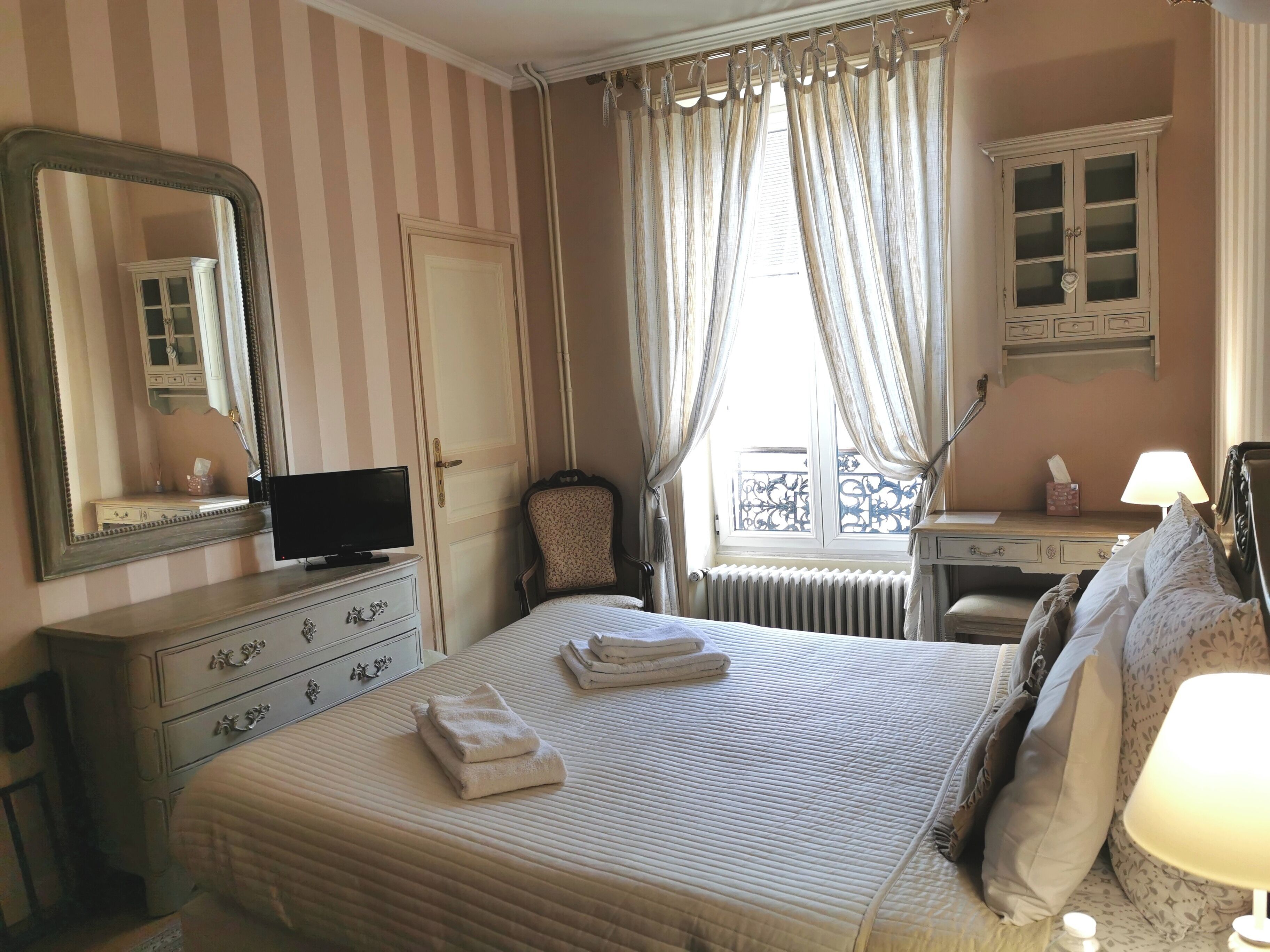 Une chambre d’hôtel comprenant un lit, une commode, un miroir, une télévision, une fenêtre avec des rideaux et un radiateur.