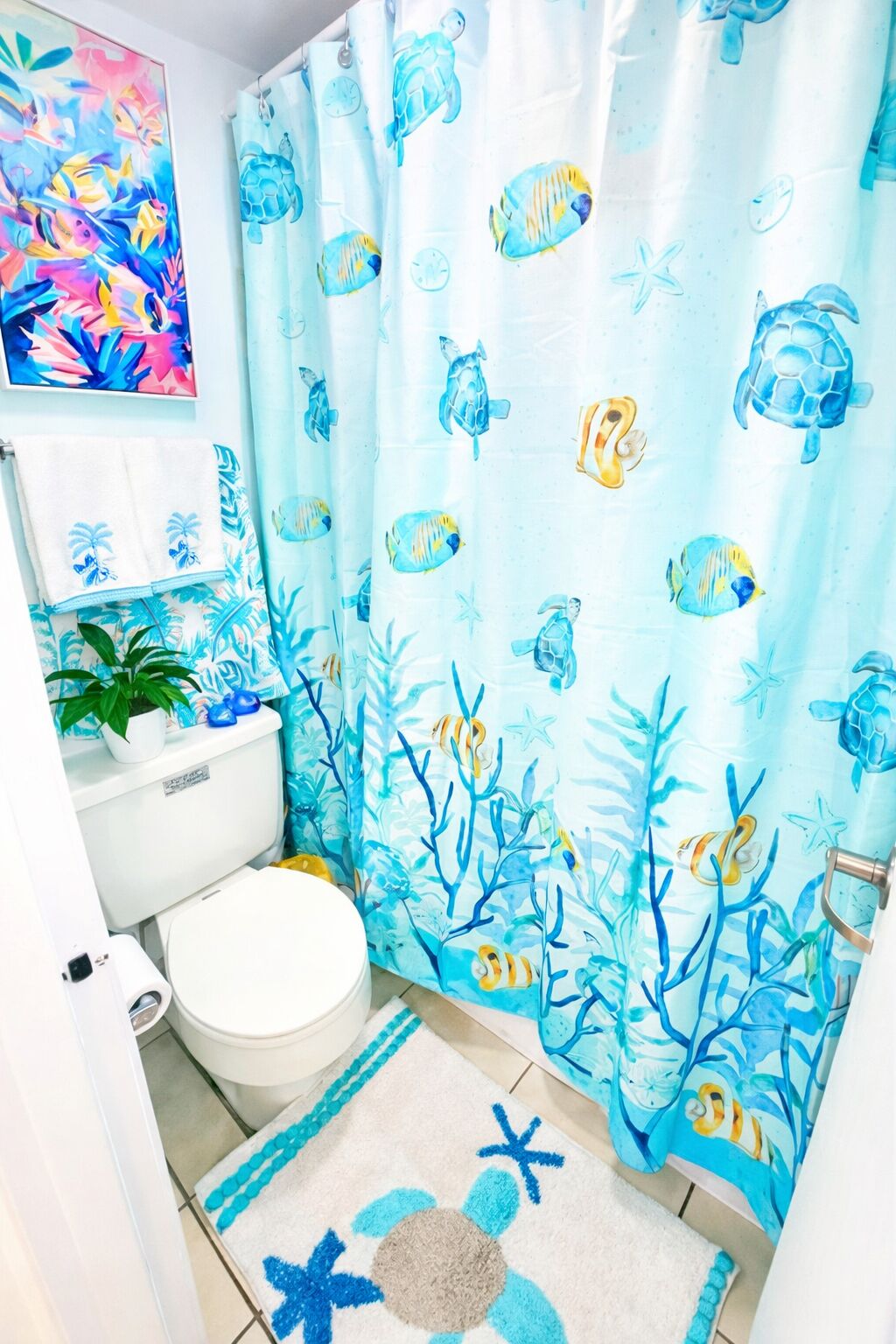 A bathroom with a light blue shower curtain featuring marine life, a toilet, a sink, and a small potted plant.