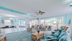 A spacious living room with a large sofa, a coffee table, and wicker chairs. There is a ceiling fan and large windows providing natural light.