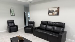 Een moderne woonkamer met een zwarte leren bank, twee zwarte leren fauteuils, een donkerhouten salontafel en een lijst met een afbeelding aan de muur.