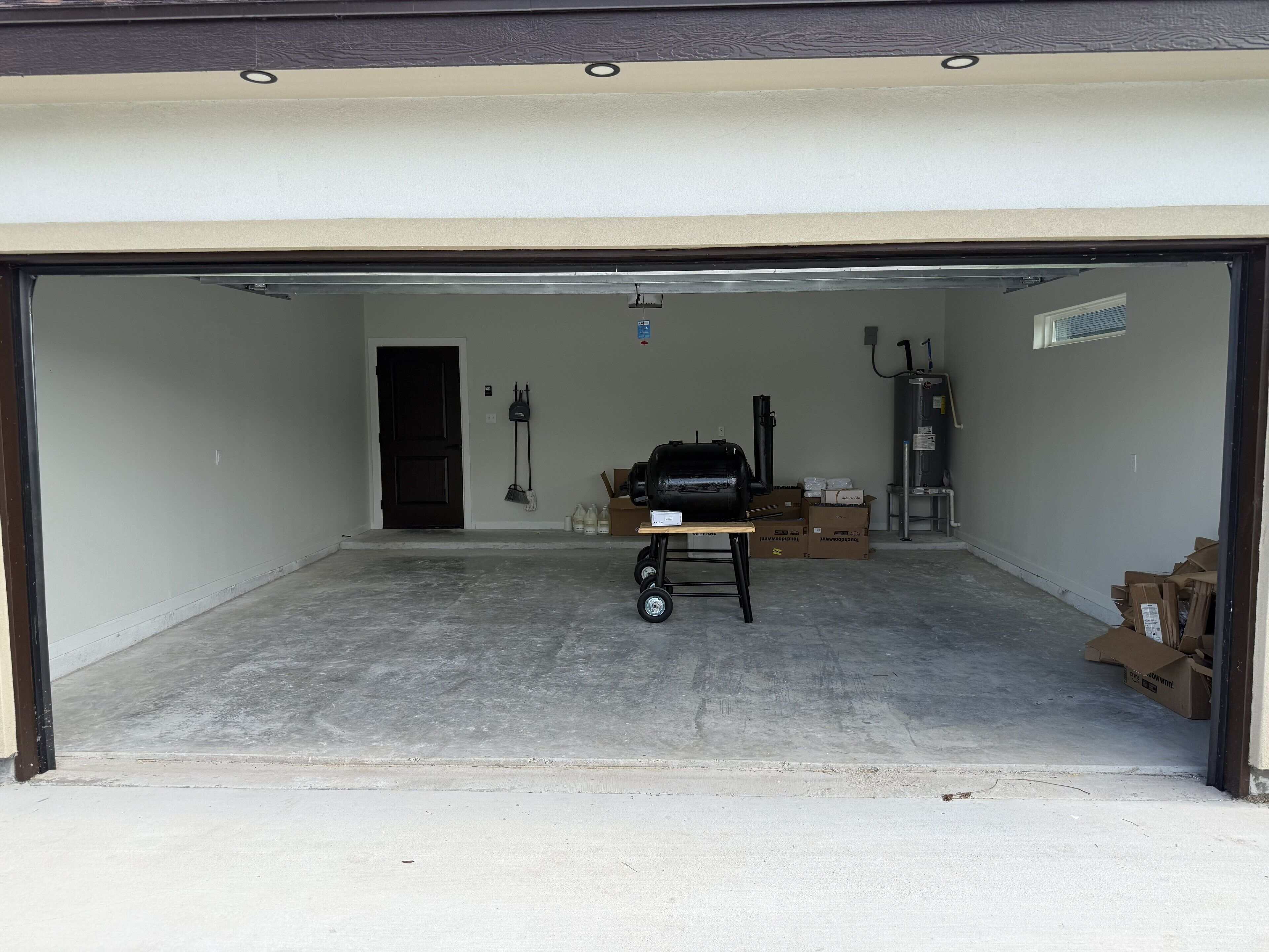 Un garage avec un plancher en béton, une porte noire et divers objets, notamment un barbecue, des cartons et un chauffe-eau.
