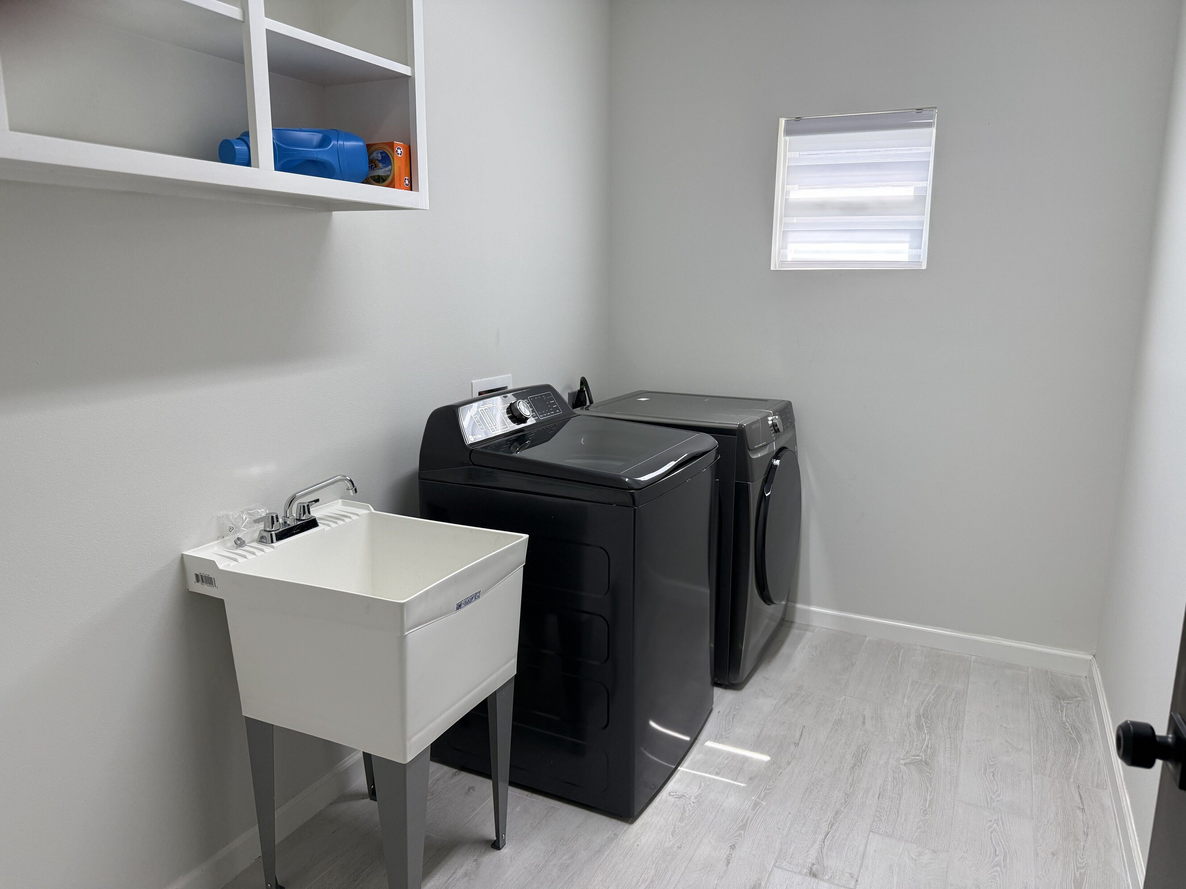 A laundry room with a white sink, two black washing machines, and a shelf with a blue container and an orange box.