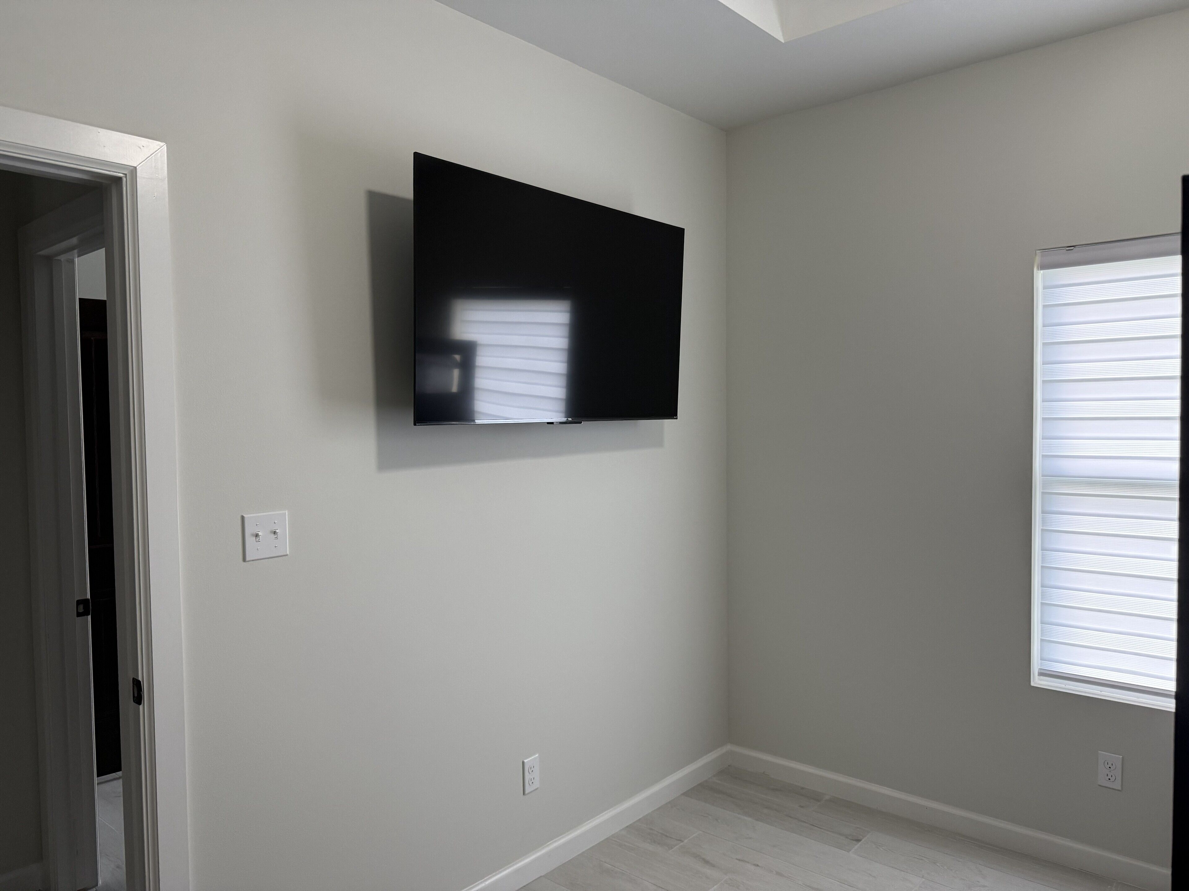 A room with a flat-screen TV mounted on the wall, a window with blinds, and a door leading to another room.