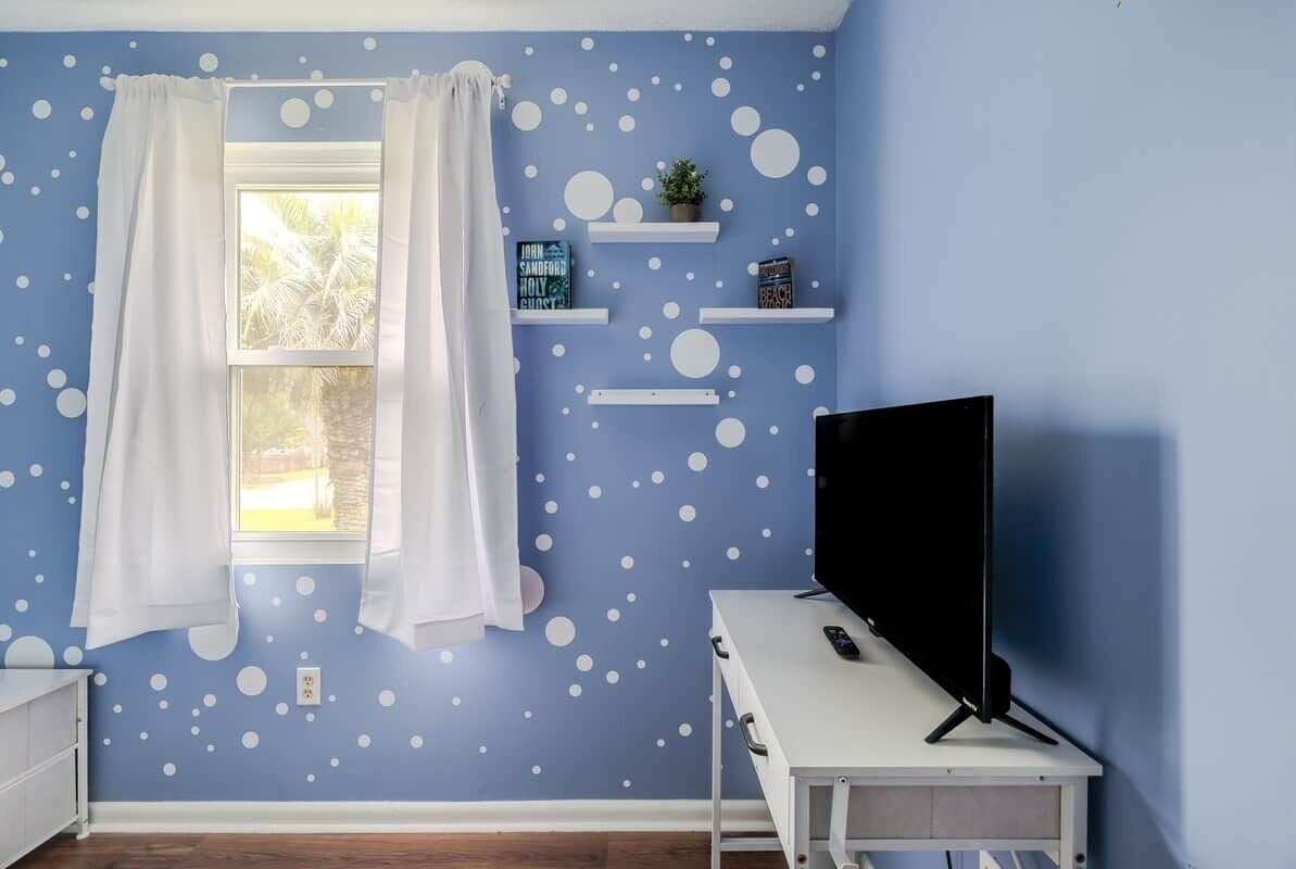 Una stanza con una parete blu, decorata con un motivo a pois bianchi, una finestra con tende bianche, una TV su un mobile bianco e scaffali con oggetti decorativi.