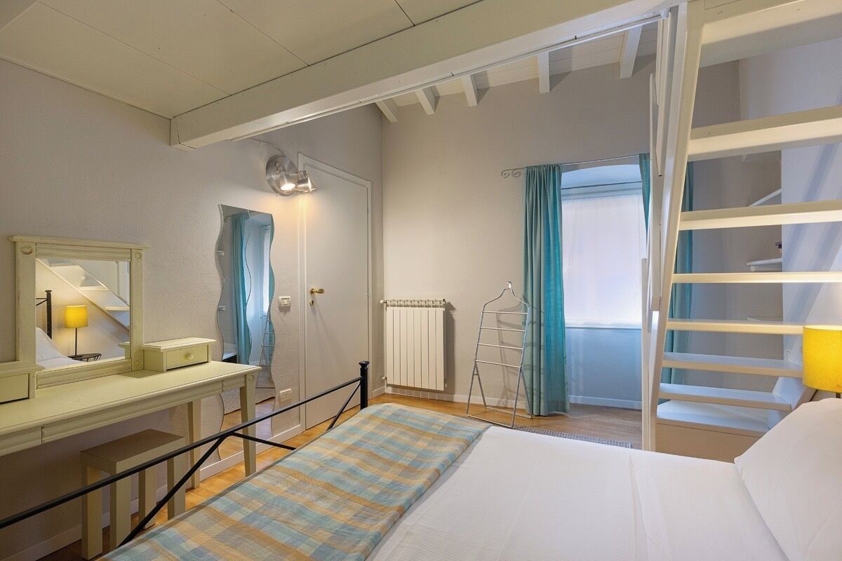 A bedroom with a bed, a desk with a lamp, a mirror, a radiator, a window with curtains, and a ladder.