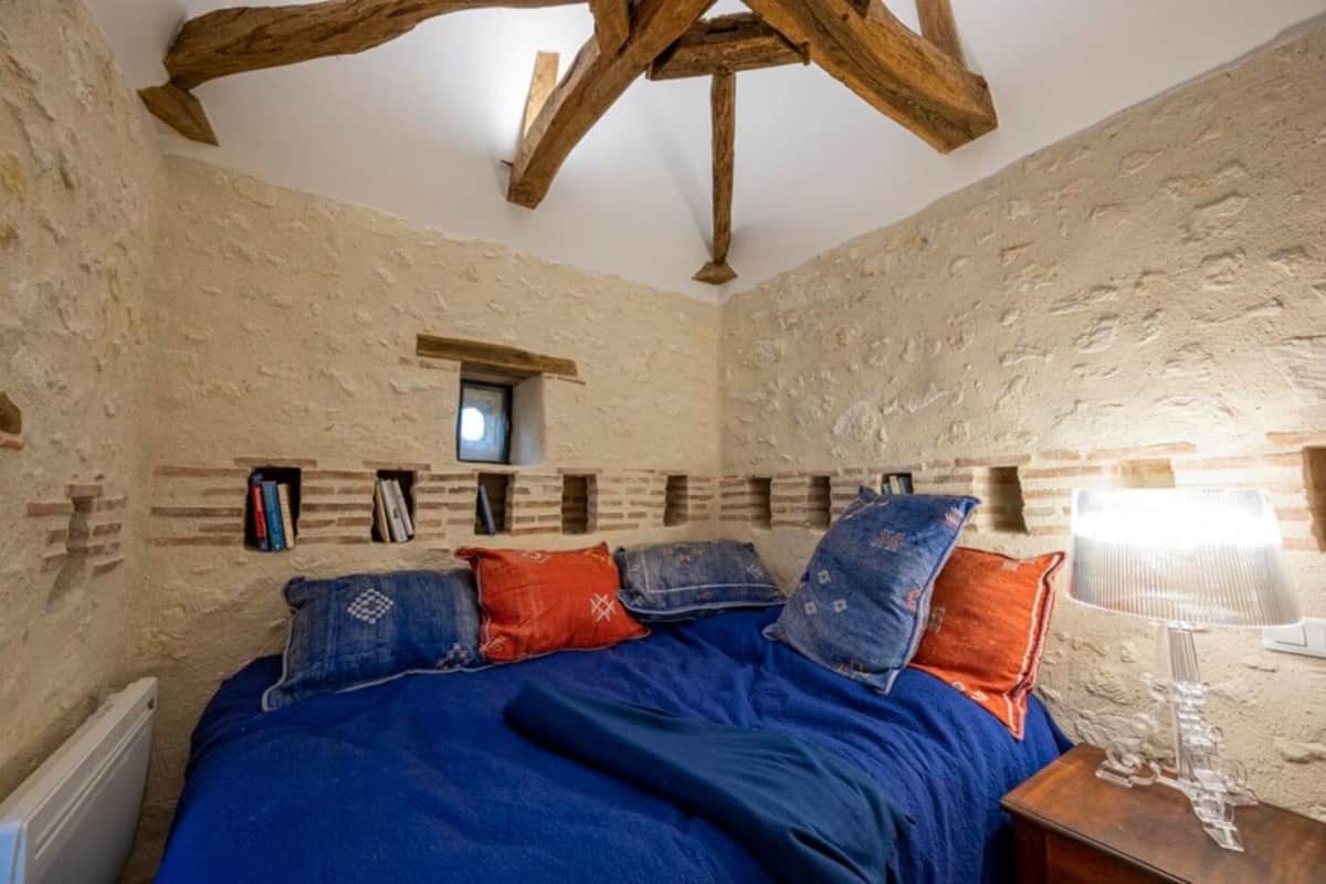 A rustic room with exposed brick walls, a wooden ceiling, and a bed with blue bedding and patterned pillows.