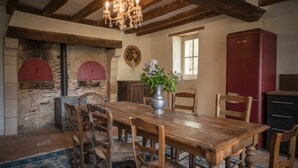 A rustic dining room with a wooden table, chairs, a chandelier, a vintage refrigerator, and a fireplace with a red arch.