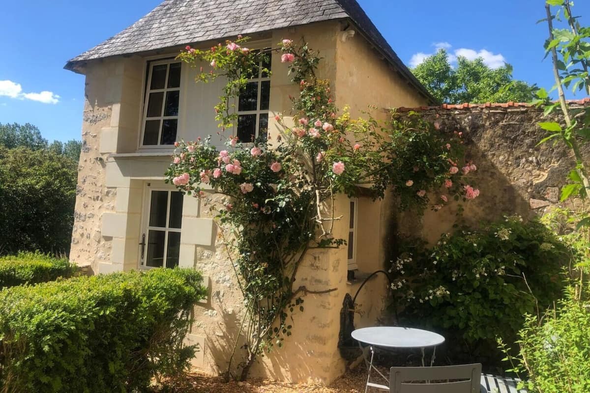 A stone house with a pitched roof, a rose bush climbing the wall, and a small round table in the garden.