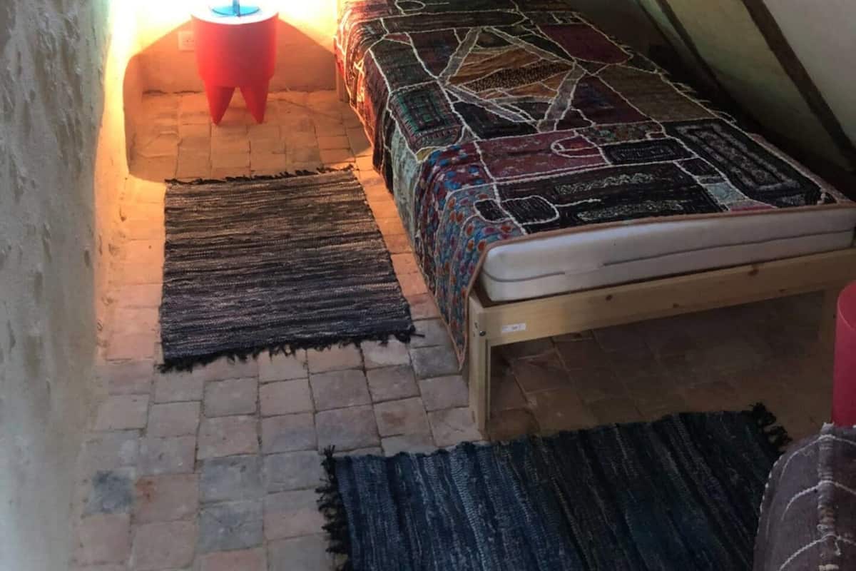A small attic bedroom with a sloped ceiling, a single bed with a patterned bedspread, a small table with a lamp, and a window providing natural light.