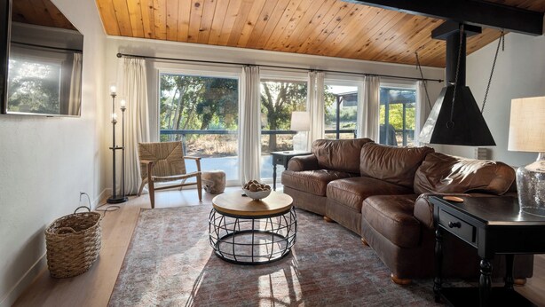 A cozy living room with a wooden ceiling, large windows, a brown leather sofa, a round coffee table, and a lamp. - Charming vacation rental steps away from River Island Golf Course (Porterville)