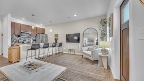A modern kitchen with stainless steel appliances, a dining area with bar stools, a living room with a sofa and a TV, and a large mirror.