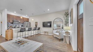 A modern kitchen with stainless steel appliances, a dining area with bar stools, a living room with a sofa and a TV, and a large mirror.