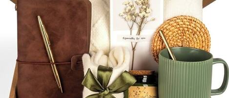 Una caja de regalo con una bolsa de cuero marrón, una taza verde, una vela, un posavasos tejido y una tarjeta con un arreglo de flores secas.