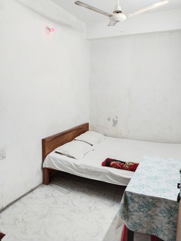 Una habitación pequeña y poco amueblada con una cama individual, cabecera de madera, ventilador de techo y una mesa con mantel estampado.