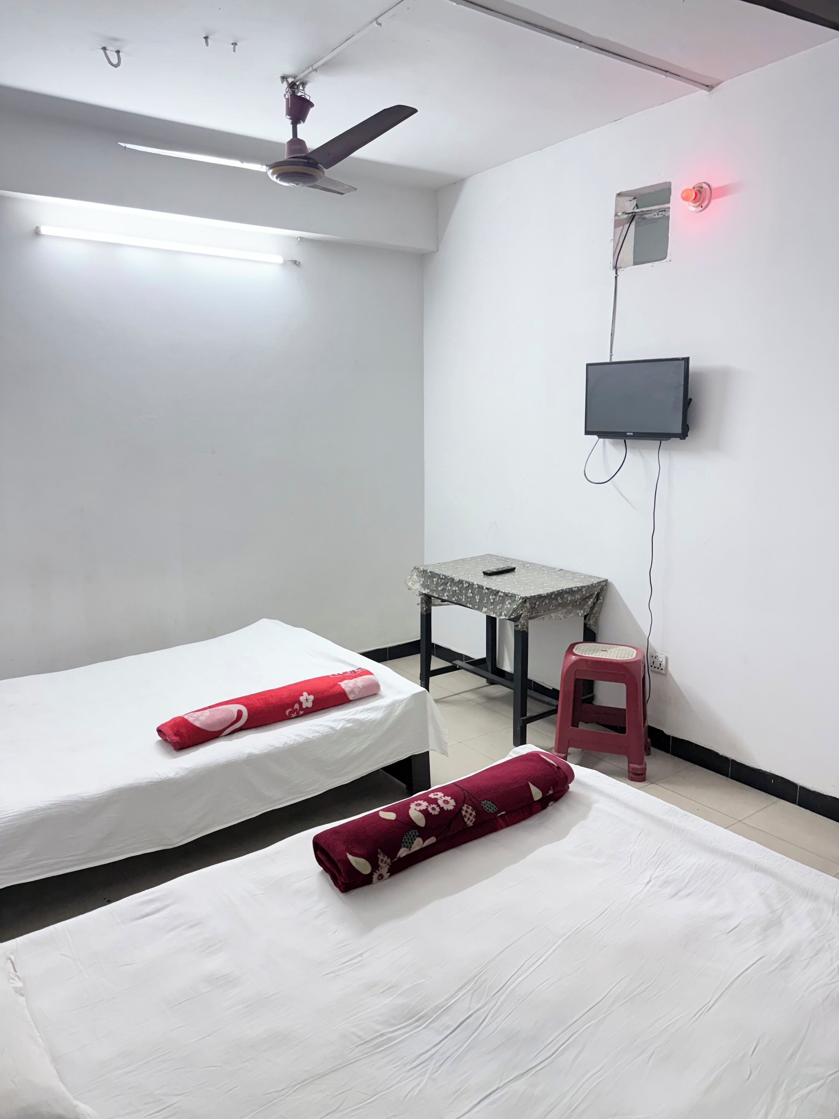 Une chambre avec deux lits, un ventilateur de plafond, un téléviseur fixé au mur, une petite table et un tabouret rouge.