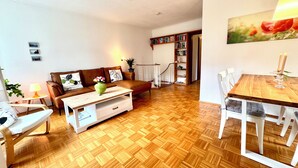 A living room with a wooden herringbone floor, a brown leather sofa, a white coffee table, and a dining area with a wooden table and white chairs. - Cozy and quiet garden vacation apartment in the heart of Dießen am Ammersee (Dießen am Ammersee)