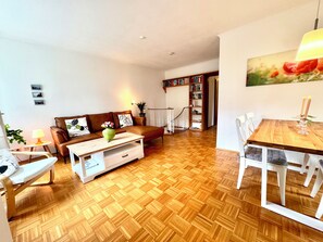 Uma sala de estar com piso em padrão herringbone de madeira, um sofá de couro marrom, uma mesa de centro branca e uma área de jantar com mesa de madeira e cadeiras brancas.
