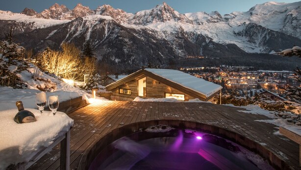 Eine Holzhütte mit einem gläsernen Whirlpool, umgeben von schneebedeckten Bergen und einer Stadt im Hintergrund.