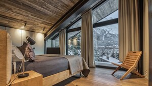 Ein Schlafzimmer mit einem großen Bett, einem Nachttisch aus Holz, einer Schreibtischlampe, einem Sessel und Blick auf verschneite Berge durch große Fenster.