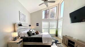 Una camera da letto con un letto, comodino, lampada, televisore, finestre e ventilatore a soffitto.