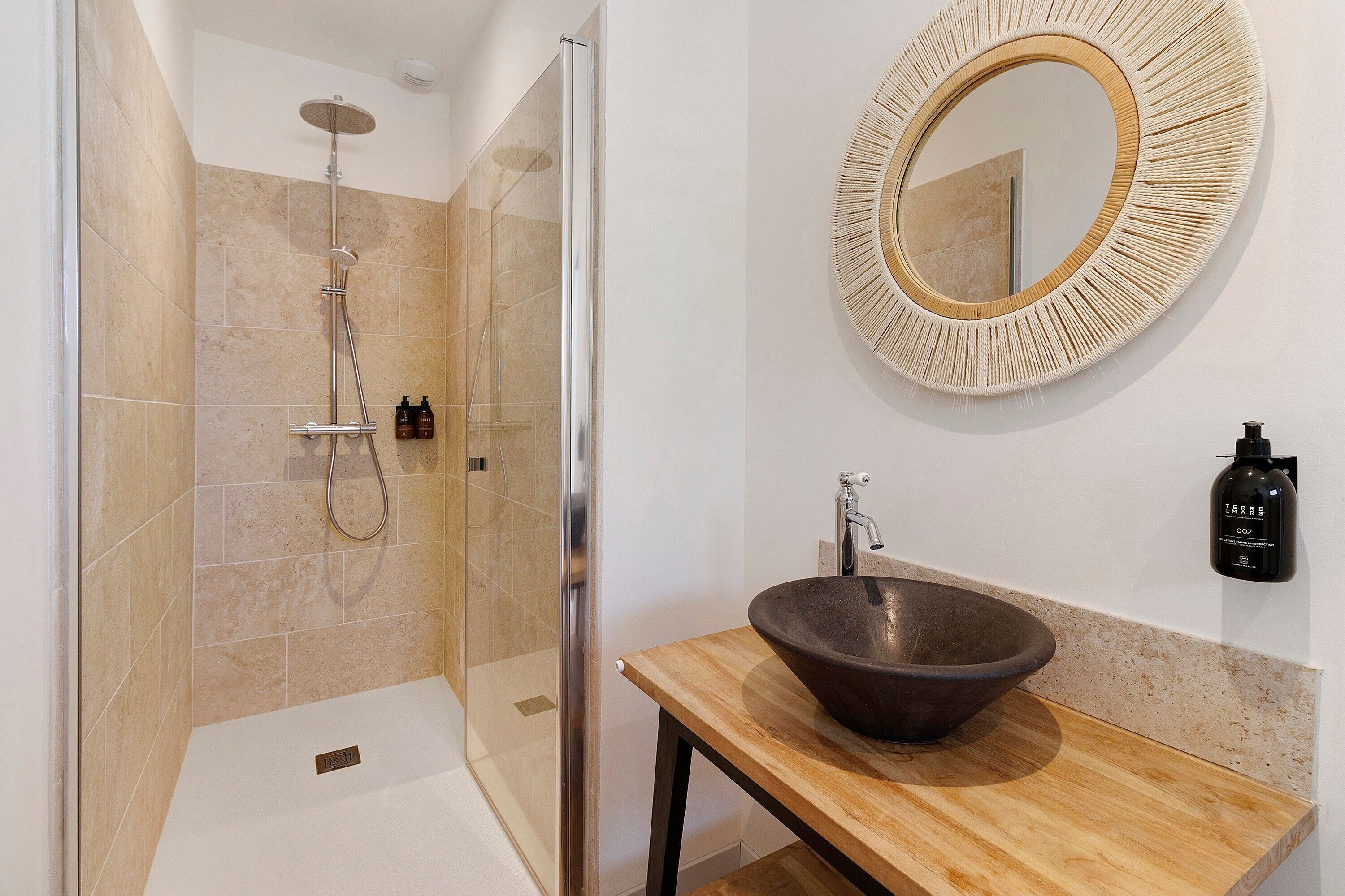 Un baño moderno con ducha de vidrio, tocador de madera con lavabo y espejo redondo con marco texturizado.