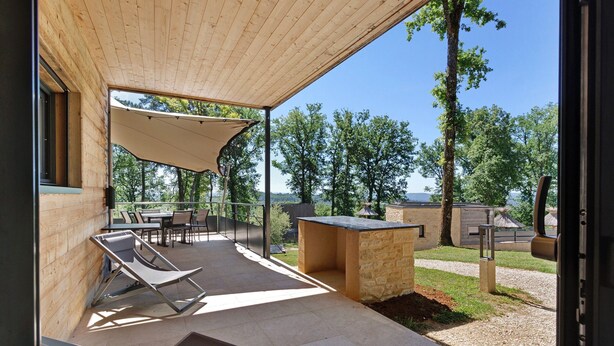 Patio esterno con tetto in legno, tavolo, sedie, zona barbecue e vista sugli alberi e il paesaggio circostanti.