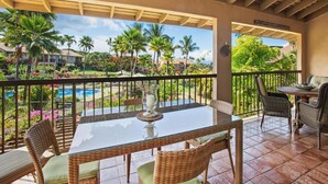 Un espace extérieur abrité, doté d’une table à plateau de verre et de chaises en osier, offrant une vue sur une piscine et une végétation luxuriante.