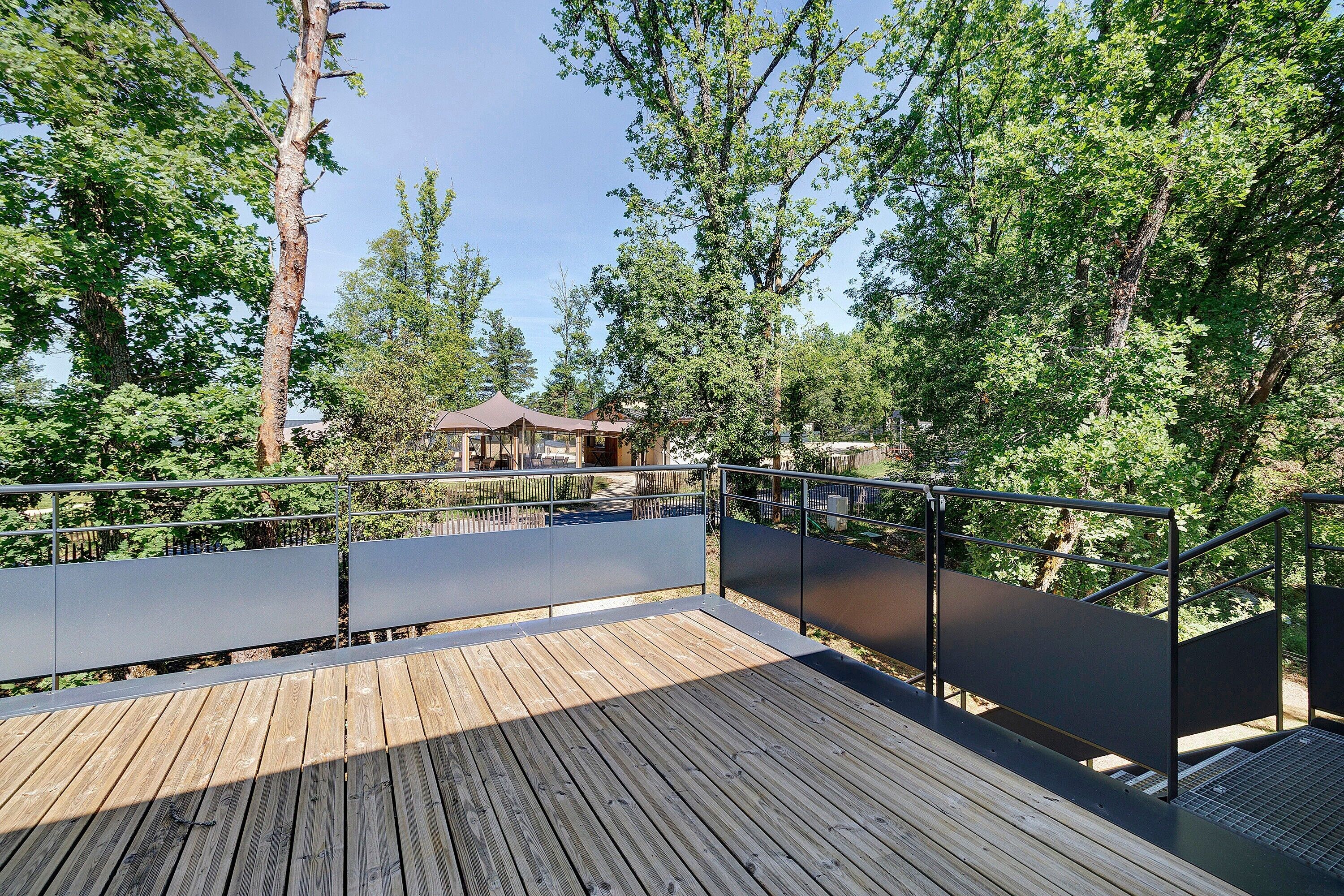 Une terrasse en bois avec des garde-corps en métal, entourée d’arbres et avec un bâtiment visible au loin.