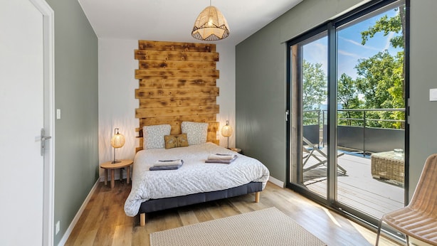 A bedroom with a wooden headboard, a large bed, a view through a sliding glass door, and a wicker chair. - Le Pech Eternel – Master Bedroom La Noyeraie (Sarlat la Canéda)