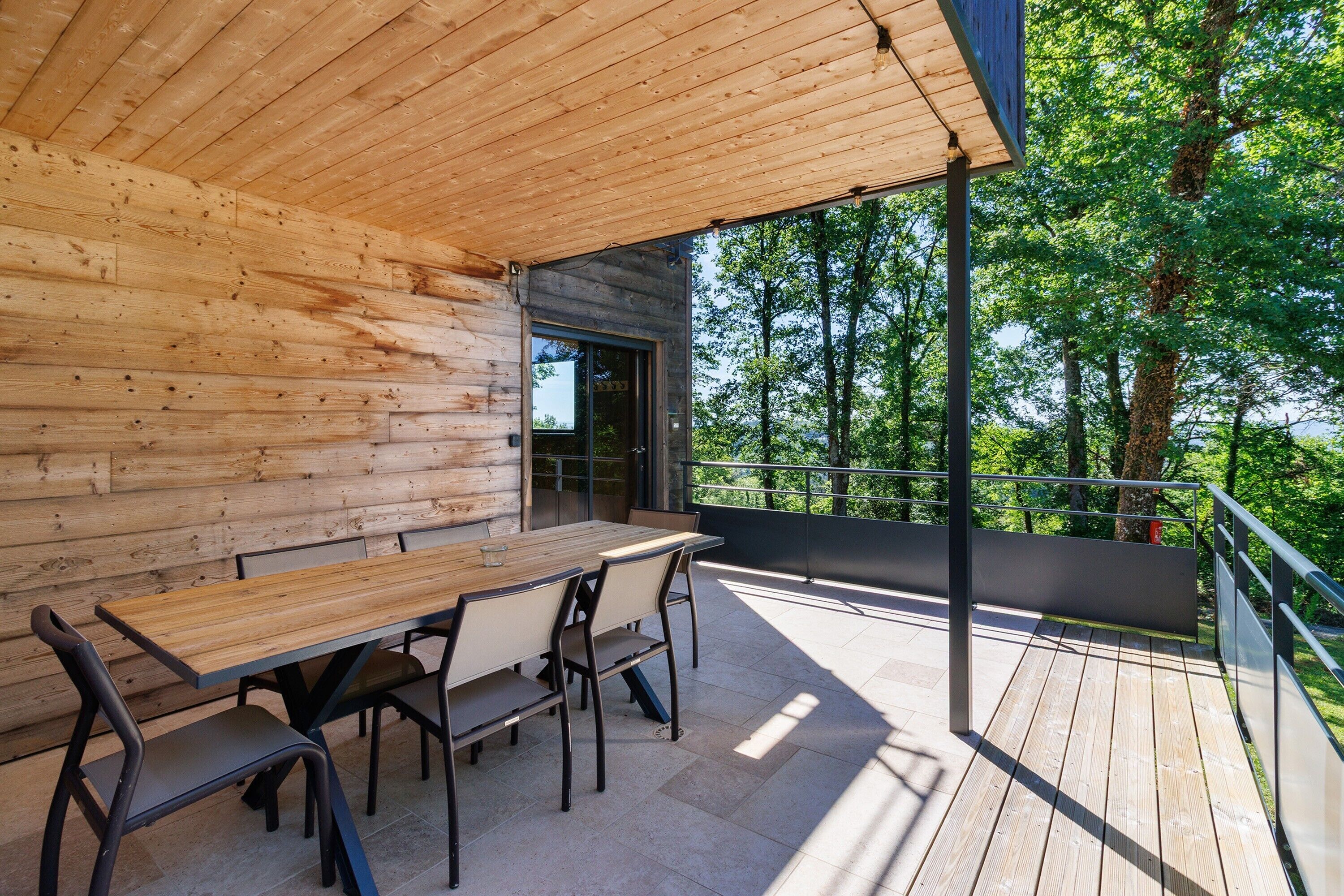 A wooden deck with a table and chairs, a balcony with a metal railing, and a view of trees.