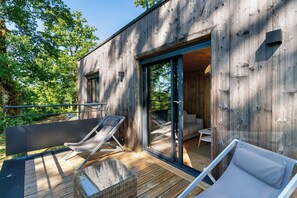 Una cabaña de madera moderna con una terraza, dos sillones de descanso y un ventanal corredizo que da a un interior con un sofá y una mesita.