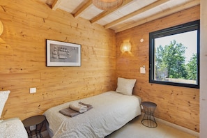 Ein Zimmer einer Holzhütte mit einem Bett, einem kleinen Tisch, einem Fenster und einer Wandleuchte.
