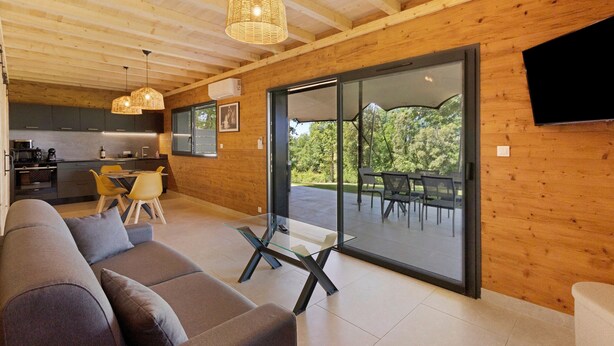 A modern interior with a sofa, dining table, and chairs, wooden walls, and a large sliding glass door to an outdoor area. - Le Pech Eternel - Lodge Spa Acacias (Sarlat la Canéda)