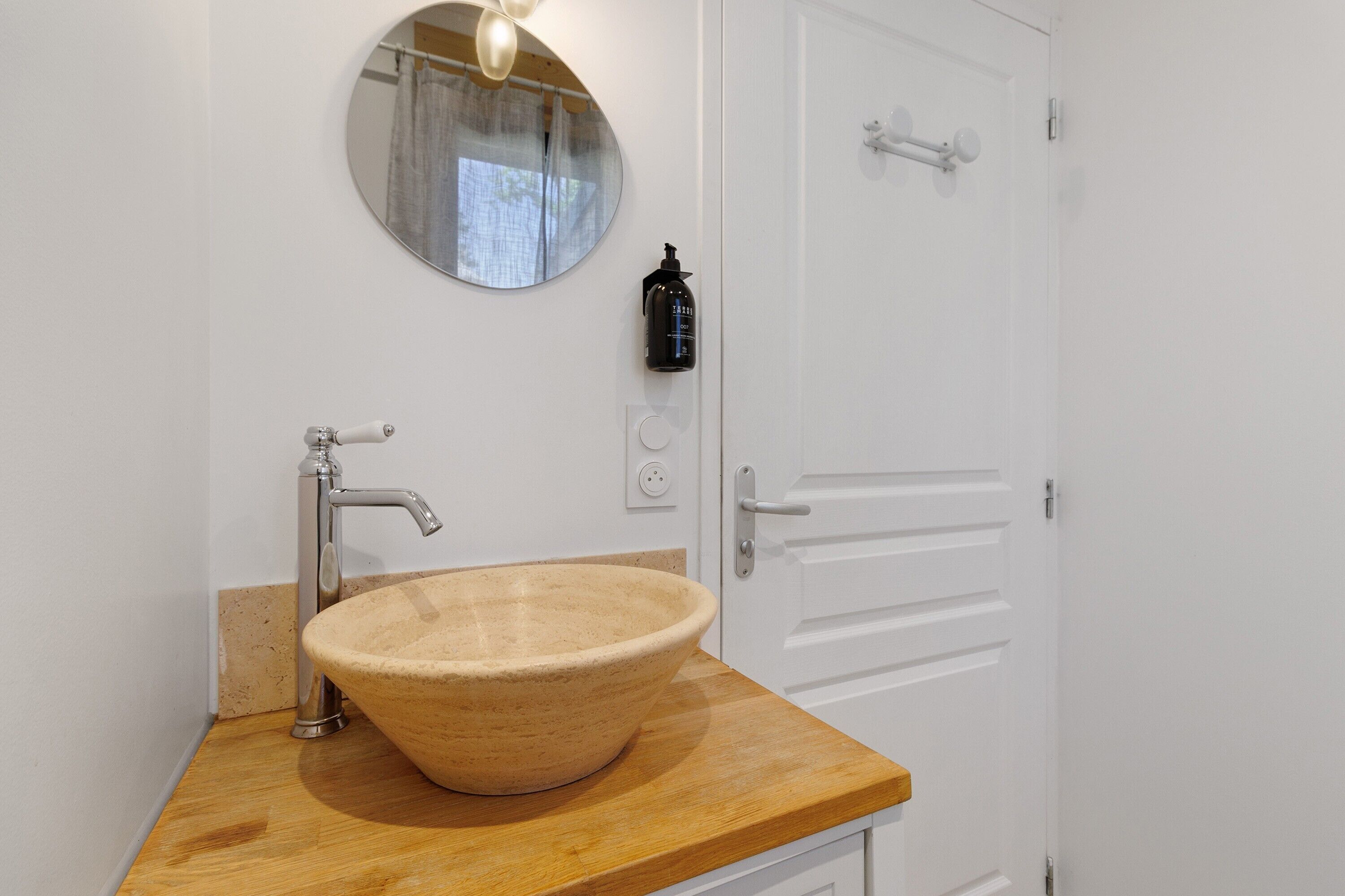 A bathroom with a wooden vanity, a round mirror, a beige ceramic sink, a modern faucet, and a door with a handle.