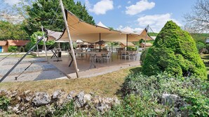 A covered outdoor seating area with tables and chairs, surrounded by greenery and a stone wall. - Holiday Home 'Les Crouquets - La Grange' with Shared Terrace, Private Garden and Wi-Fi (Jayac)