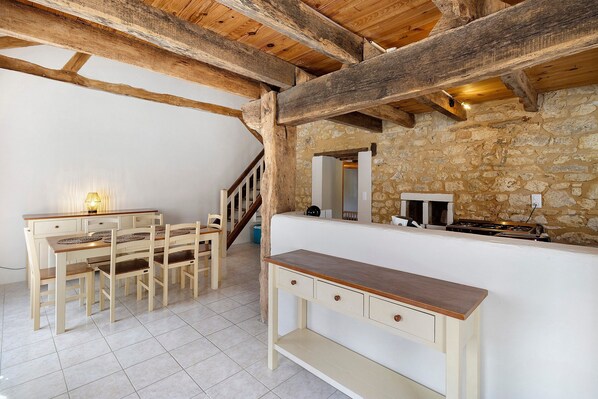 A rustic interior with exposed wooden beams, a stone wall, a wooden dining set, and a kitchenette.