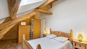 A bedroom with a sloped ceiling, wooden beams, a bed with white bedding, a nightstand with a lamp, and a wooden wardrobe. - Les Crouquets - Le cerisier (Jayac)