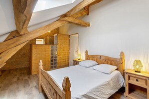 Ein Schlafzimmer mit geneigter Decke, Holzbalken, einem Bett mit weißer Bettwäsche, einem Nachttisch mit Lampe und einem Holzschrank.