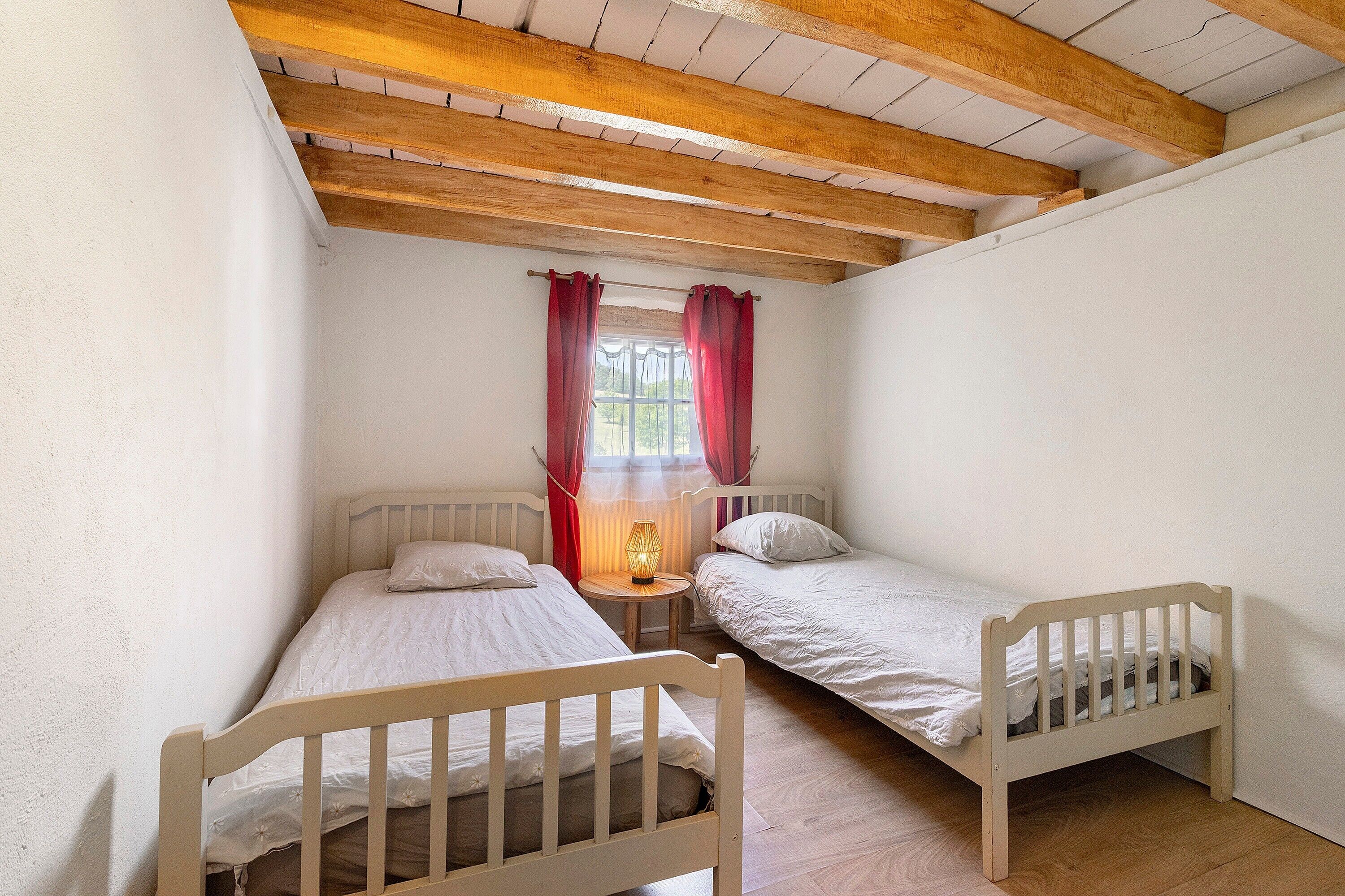 Quarto com duas camas de solteiro, vigas de madeira no teto, uma mesa pequena com abajur e uma janela com cortinas vermelhas.