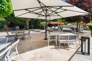 An outdoor patio with tables and chairs, a large umbrella, and a pool area in the background.